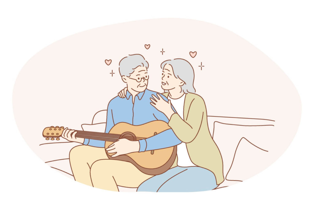 Old couple sitting down on a sofa while she hugs him from behind as he plays the guitar